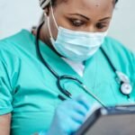 Nurse with mask and gloves reviewing documents on clipboard in a clinic setting.